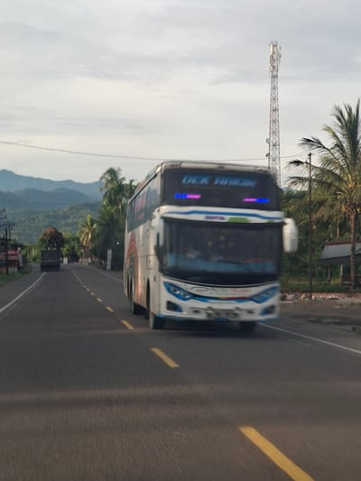 An over-the-shoulder view captures a blurred white and blue bus on a two-lane asphalt road. The bus, oriented slightly to the right, displays a rainbow graphic on its side and illuminated blue text that appears to read "DEK HAKIM" above the windshield. Yellow lane markers are visible on the road. In the background, a cell tower stands tall against a hazy sky. To the right of the bus, a palm tree and a small, rustic shelter are present. On the left side of the road, a dark-colored truck is partially visible, and beyond it, rolling hills with dense foliage are seen. The overall scene suggests a rural or semi-rural setting in Meukokuthang, Indonesia, with the blurred motion indicating the vehicle is in transit.