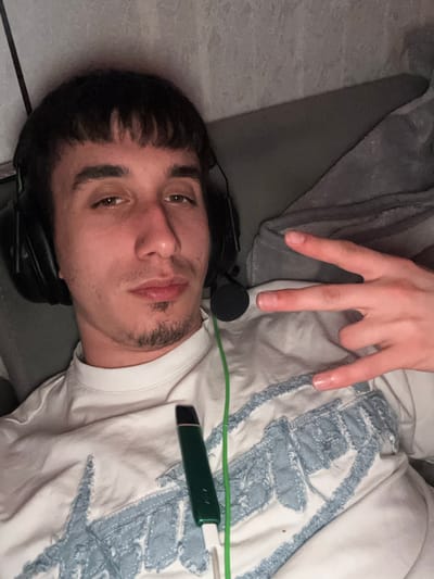 The scene is set indoors, within the city of Vetrino, Bulgaria. A single male individual is depicted in a medium close-up, facing the camera directly. He possesses dark, short hair styled with a straight fringe across the forehead and light stubble on his chin and upper lip forming a goatee. He wears black over-ear headphones with an attached microphone boom, which is positioned to his left. A thin, bright green cable extends downwards from the headphones across his chest. He is dressed in a light-colored (possibly off-white or cream) long-sleeved shirt featuring a distressed light blue fabric design on the front. His left hand is raised to chest level, forming a gesture with the index and pinky fingers extended and the thumb, middle, and ring fingers curled. Resting on his shirt is a dark green, pen-shaped device, likely an e-cigarette or vape pen, which is connected to a short white charging cable. This white cable is draped over the green headphone cable. The individual appears to be reclining against a grey upholstered surface, possibly a sofa or bed, with a grey textured wall visible in the upper background.