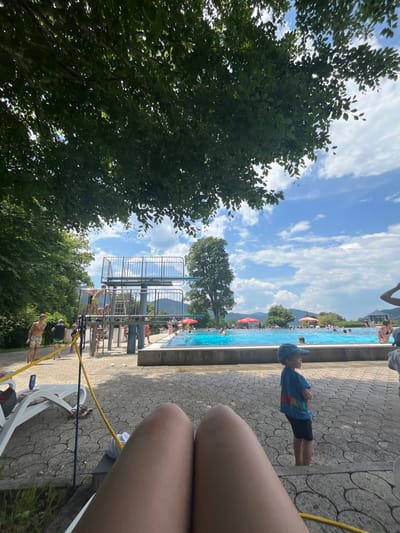 The image depicts a sunny day at an outdoor swimming pool facility in Oberammergau, Germany, likely the "Wellenbad". In the foreground, a person's legs are visible, suggesting a first-person perspective from a lounge chair. The background features a large, bright blue swimming pool with several people swimming and relaxing. A diving platform with multiple levels is present to the left of the pool, with a few individuals preparing to jump or observing. The poolside area is paved with a patterned stone surface, and several lounge chairs are scattered around. Red umbrellas provide shade for some areas. Lush green trees and foliage surround the pool area, and in the distance, mountains are visible under a partly cloudy blue sky. A young boy wearing a blue cap and t-shirt stands near the pool's edge, facing away from the camera.