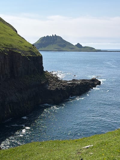 The media file shows a landscape view of the ocean and cliffs in Gásadalur, Faroe Islands. The scene is dominated by rugged, grassy cliffs plunging into a deep blue, rippling ocean. In the foreground, a dark, jagged rock formation juts out into the sea, with waves crashing against its base, creating white foam and glittering highlights on the water's surface. The cliffs are covered in vibrant green grass, dotted with small yellow flowers and some darker patches of rock. In the distance, more imposing, serrated mountains rise from the sea, partially shrouded in soft, hazy light under a pale blue sky with scattered white clouds. The overall impression is one of wild, natural beauty and a sense of vastness. The sun appears to be shining, creating bright reflections on the water, suggesting it is likely daytime. There is no text visible in the image.