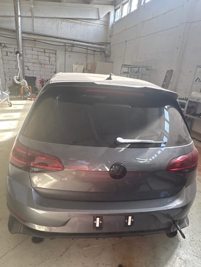 The image shows the rear view of a dark gray Volkswagen Golf R, parked indoors. The car is in what appears to be a workshop or garage. To the left of the car, a large metal exhaust pipe system is visible, curving upwards. Further to the left, a yellow air compressor machine is present. The background consists of brick walls and shelving units. Sunlight streams in from windows at the top of the frame, casting rectangular reflections on the car's rear window and body. There are no people visible in the image. The scene is located in Schkopau, Germany.