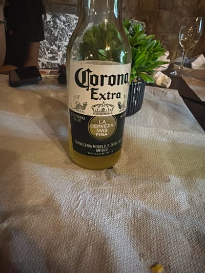 A close-up, eye-level shot focuses on a bottle of Corona Extra beer positioned on a textured, light-colored tablecloth. The bottle is partially filled with a golden liquid, and the label is clearly visible, featuring the brand name "Corona Extra" in bold black lettering, along with other text in Spanish and English indicating origin and volume. To the right of the bottle, a small, decorative green plant in a black planter is visible. Further to the right, a partially obscured wine glass with a light-colored liquid is present. In the background, a person's dark-clad shoulder and arm are visible, along with a portion of a wall with a textured, abstract pattern. The overall setting suggests an indoor environment, possibly a restaurant or bar. There is no information or visual data in the image to indicate the location being Kermen, Bulgaria.
