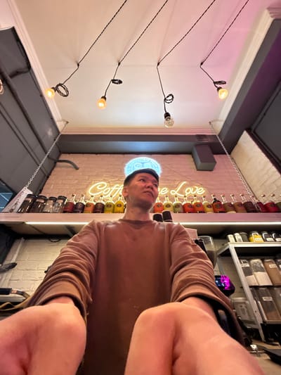 In the city of Veshki, Russia, an individual is positioned centrally in the frame, looking upwards and to the right. They are wearing a brown sweatshirt and a dark cap. The background features a brick wall with a neon sign that reads "Coffee Love." Above the wall, a shelf displays an array of bottles and jars. The ceiling is adorned with multiple hanging light fixtures, each with a single exposed bulb. The scene appears to be an interior space, possibly a coffee shop or a similar establishment.