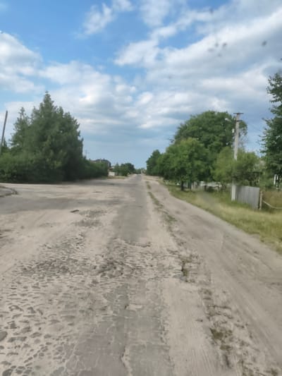 The image depicts a dirt road in the city of Bohushi, Ukraine, under a partly cloudy blue sky. The road is heavily potholed and appears to be in disrepair, with loose gravel and dirt. Trees line both sides of the road, with dense foliage on the left and a mix of trees and fences on the right. In the distance, houses and utility poles are visible, suggesting a rural or semi-rural residential area. The overall scene conveys a sense of an unpaved, weathered rural landscape.
