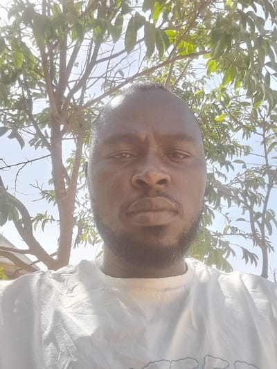 The specific location within Barakin Acha, Nigeria, is not discernible from the visual evidence. The image depicts an adult male, with dark skin, short dark hair, and a trimmed beard, looking directly forward with a neutral expression. He is wearing a light-colored, likely white, crew-neck t-shirt with visible creases. The background features tree branches with green leaves, illuminated by bright daylight, against a clear sky, indicating an outdoor, sunny environment.