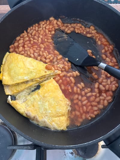 This image shows a close-up view of a black frying pan on a stovetop. The pan contains baked beans in a tomato sauce and a folded omelet. A black spatula is in the pan, partially submerged in the beans, appearing to be in the process of serving or mixing them. The beans are a light brown color and appear plump and cooked. The omelet is golden yellow and folded into several layers. The scene is indoors, likely in a kitchen, as suggested by the presence of the stovetop and the cooking food. There are no people visible in the image. The lighting appears to be natural or overhead room lighting. No text is visible in the image. The overall impression is of a simple, home-cooked meal being prepared or served.