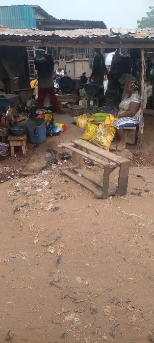 The image depicts an open-air workshop or market stall in Ilogbo Eremi, Nigeria, under an overcast sky. The foreground features a reddish-brown dirt ground scattered with small white debris and dark fragments, where a wooden frame or pallet lies on its side.
Under a weathered corrugated metal roof supported by wooden posts, several individuals and various objects are arranged. To the right, a woman sits on a low wooden stool. She wears a patterned light-colored top, a striped wrapper, and a headscarf, her bare feet resting near a sandal. Next to her are several yellow sacks, some green plastic basins, and a yellow container.
Towards the center and left under the shelter, an active work area is visible. A person wearing a dark top and red trousers stands with their back to the camera, appearing to work on machinery. Another individual in a dark top is similarly engaged further right. The area contains an array of items including engines, mechanical parts, metal bowls, a blue plastic barrel, and various tools and scrap materials. A large black circular object, possibly a flywheel, rests on a wooden support.
In the background, beyond the immediate structure, other buildings are visible, including one with a light blue corrugated metal siding or roof, and another with a barred window.