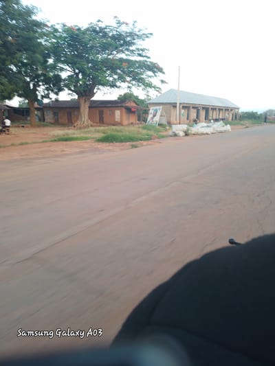 The image shows a street scene in Jibilik, Nigeria. On the left side of the road, there are some mud brick buildings with thatched roofs, and a large tree with green foliage and orange flowers. A person in a white shirt and dark shorts is seated on a bench in front of one of the buildings. Further down the road, there is a longer building with a metal roof and a colonnaded facade. Bags of what appear to be produce are stacked in front of this building. Several people are visible in front of and inside the building, and some motorcycles are parked nearby. The road itself is paved with asphalt and shows signs of wear. The sky is overcast. The bottom of the image displays the text "Samsung Galaxy A03", indicating it is a captured image from a Samsung Galaxy A03 device.