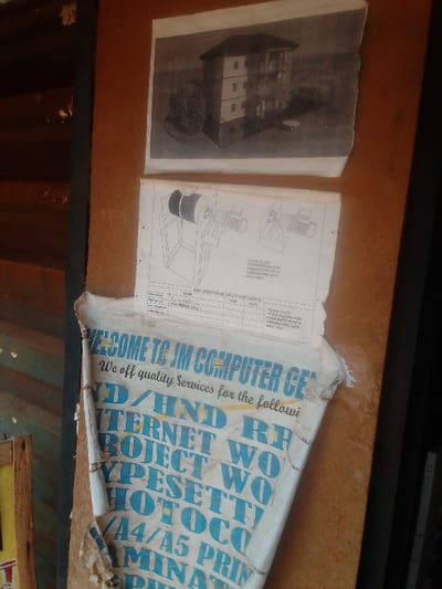 The image depicts a wooden surface with three pieces of paper pinned to it. The topmost paper is a black and white photograph of a three-story building with a green bush in front of it. The middle paper is a diagram of a piece of machinery with text that reads "Scale: 1:100, Design, Implementation and Fabrication of a Laboratory Size Ball Mill." The bottom paper is a torn, faded, and yellowed advertisement for a business called "JM Computer Center" in blue and yellow lettering. The advertisement advertises services including "D/HD," "Internet RF," "Project Work," "Typeset Work," "Hot Occ," "A4/A5 Print," and "Laminate." The location is Auchi, Nigeria, based on coordinates (latitude: 7.062, longitude: 6.273).