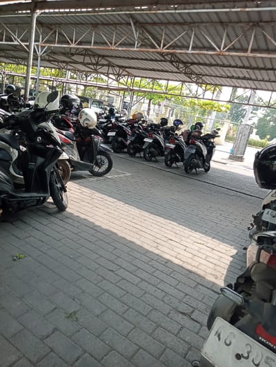 The scene depicts a motorcycle parking area in Sukomoro, Indonesia. Numerous two-wheeled scooters and motorcycles, primarily black, grey, white, and red, are parked in multiple rows on a grey interlocking brick pavement. Several helmets, including a white one with a floral pattern and multiple plain black or white helmets, are resting on the parked vehicles. The parking area is covered by an open-sided structure featuring a corrugated metal roof supported by white, visibly rusted, metal trusses and vertical posts. Sunlight illuminates the area from the left and front, casting distinct shadows of the vehicles and the structure onto the paved ground. In the background, beyond the parking facility, green foliage, a fence, and portions of distant structures are visible. No individuals are present. License plates visible on some motorcycles include "AG 305" on a Honda brand motorcycle in the right foreground, and other plates beginning with "AG".