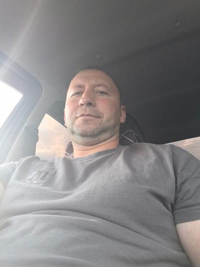The image shows a man with short hair and a beard sitting in a car. He is wearing a grey t-shirt with a logo on the left side of his chest. The sun is shining through the car window, creating a lens flare effect on the left side of the image. The man is looking directly at the camera. The background of the image is blurry, but it appears to be a road or a street. There are no other people or objects visible in the image. The man is not interacting with anyone or anything. The scene is likely taking place during the daytime.