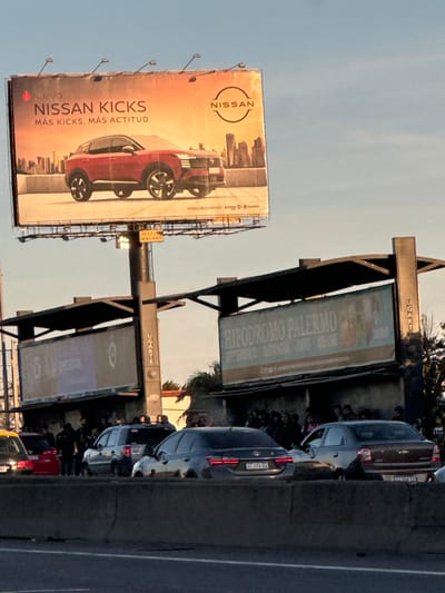 The image captures a roadside scene in Olivos, Argentina, featuring several prominent elements. There is a large billboard advertising the "Nissan Kicks," another advertising "Hipódromo Palermo" billboards with "Ugarte" signs on top, and traffic on a road adjacent to the signs. The lighting suggests late afternoon, and the sky is clear. There are several cars, and also what appear to be people walking or standing nearby, potentially in a line or crowd.