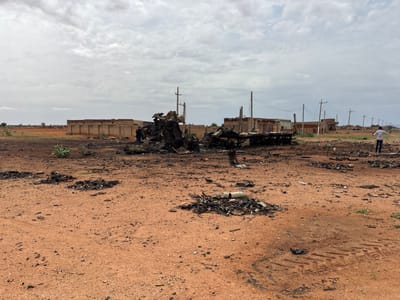 In Um Laba, Sudan, the scene depicts the aftermath of a significant incident, with two individuals present. One person, dressed in dark clothing, stands near a heavily damaged vehicle in the mid-ground. Further to the right, another individual in a white shirt and dark pants gestures with their arms outstretched. The environment is an open, arid area with red soil, scattered debris, and remnants of burnt structures in the background. The foreground is littered with ash, burnt material, and tire tracks, suggesting recent destruction. A partially intact building with a long, low facade is visible behind the primary wreckage, along with utility poles and power lines extending into the distance. The sky is overcast with clouds.