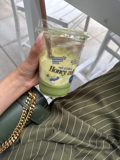 A person's hand with light-colored, manicured nails holds a clear plastic cup filled with a green, creamy beverage. Condensation beads on the outside of the cup. The cup has a blue label with white text that reads "Your everyday handmade break" and "Honey rev cafe & bakery". Below "Honey rev", there is a small drawing of a bee. The person is wearing a dark green, pinstriped shirt. A dark green purse with a thick gold chain strap is visible next to the cup and shirt. The background appears to be an indoor or outdoor setting with bright, natural light, suggesting daytime. The metallic railings and tiled flooring visible in the background suggest a public area like a mall, cafe, or beachside promenade. The overall mood is casual and relaxed, perhaps a moment of enjoying a refreshment. The text indicates the beverage is from "Honey rev cafe & bakery". No specific time of day or weather conditions are explicitly mentioned beyond the bright lighting.