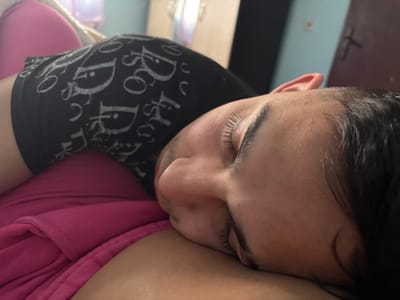 An overhead-angle image depicts an adult male asleep or resting, positioned on a pink fabric surface within an indoor residential setting in Gorna Rositsa, Bulgaria. The individual is shown from the upper chest to the head, with eyes closed and face turned to the left. He has short, dark hair and visible stubble on his jawline. He is wearing a black short-sleeved garment or underwear patterned with repeating white stylized "D" logos, resembling the "Dior" emblem. The pink fabric beneath him, possibly a blanket, pillow, or another person's clothing, is folded and textured. The background is out of focus, revealing a dark brown door with a metal handle to the upper right, and a light blue wall section above it. Minimal natural or ambient artificial light illuminates the scene. No other individuals or active interactions are directly visible.