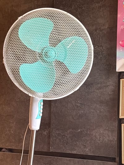 A close-up shot shows a white oscillating fan with light blue blades. The fan is positioned against a dark, textured background that appears to be a wall. The fan's grill is made of fine white mesh with a diamond pattern. The blades are wide and triangular, with a lighter blue accent near the center. The fan's motor housing is white with light blue accents and has several small, round buttons on its side. A thin white cord is visible extending from the motor housing and disappearing out of frame to the right. The overall impression is of an indoor setting, possibly a room, with a focus on the fan as the main subject. There is no indication of the time of day or weather. No visible text is present in the image. The context provided indicates the location as Yanevo, Russia, but this cannot be visually confirmed from the image itself.