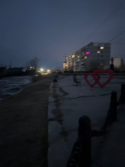 The image, likely taken in the town of Zhatay, Russia, shows a dark, twilight scene with a tall, multi-story building visible in the background. Many windows in the building are lit up. In the foreground, there is a metal structure consisting of two interconnected hearts. A pedestrian walkway or sidewalk, possibly wet from rain or snow, is visible. A low, dark barrier runs along the edge of the sidewalk, and beyond that is a grassy area that appears to be snow-covered in places. Street lights are positioned along the walkway, providing some illumination. The overall atmosphere is dim and urban.