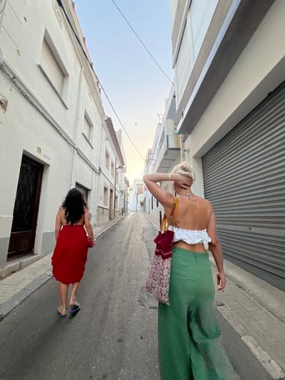 Two women walk down a narrow, paved street in L'Escala, Spain. The woman in the foreground, with blonde hair pulled up, wears a white cropped top and long green pants. She carries a patterned tote bag over her right shoulder and rests her left hand on the back of her head. She has tattoos on her back and left arm. The woman behind her has dark, curly hair and wears a long, flowing red dress. She carries a small red clutch in her right hand and wears flip-flops. The street is lined with white buildings, some with shuttered windows and others with balconies. A large gray metal roll-up door is visible on the right side of the image. Power lines run overhead against a pale blue sky. The image appears to have been taken in the late afternoon or early evening, as evidenced by the soft, diffused light.