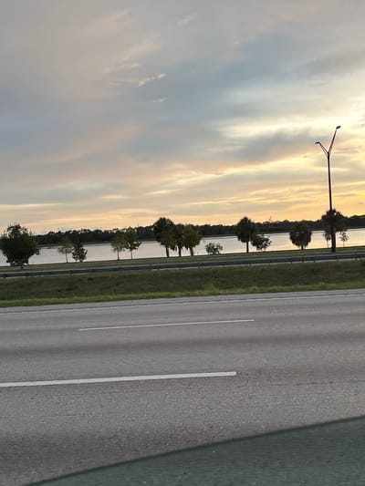 An eye-level, medium shot captures a roadside view of a lake and a strip of trees, with a highway in the foreground. The sky is filled with soft, muted clouds, tinged with pale yellow and orange, indicating either dawn or dusk. A single streetlamp stands tall on the right side of the frame, its pole extending upwards into the sky. The highway, paved with asphalt, is marked with white lines, suggesting it is a multi-lane road. The water of the lake is calm and reflects the muted colors of the sky. A line of trees separates the lake from the highway, their foliage a mixture of green and darker shades. The overall scene conveys a sense of tranquility, characteristic of a suburban or natural area bordering a major road. The location is identified as being in the city of Sarasota, United States.