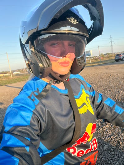 A person wearing a black motorcycle helmet and a blue and black racing-style jacket with "Red Bull" and "Fox" logos is seen in an outdoor setting. The person has a nasal cannula in their nostrils. The ground appears to be gravel or small stones. In the background, there is a road, grassy area, and utility poles. There is a building in the distance. The location may be Азьмушкино, Russia, based on the instructions.