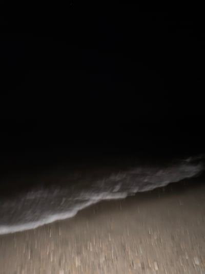 A nighttime photograph captures a section of the beach in Torreilles, France. The image is dominated by darkness, with the upper portion entirely black. The lower third of the frame shows the shoreline where a wave is breaking. The wave is depicted as a foamy white line, suggesting motion blur due to the low light conditions and possibly a long exposure. The beach itself is visible as a textured area of sand or small pebbles, appearing blurred and slightly illuminated from an unseen source. The scene conveys a sense of quiet and natural beauty in the dark.