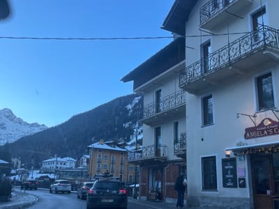 The image captures a street scene in a mountainous town, identified as La Thuile, Italy, during what appears to be early morning or late afternoon due to the soft, cool light and clear blue sky. Snow is visible on the mountains and sporadically on the ground and rooftops, indicating cold or winter weather.

On the right side of the frame, a multi-story building with light-colored facades features several ornate black wrought-iron balconies. The ground floor houses "ANGELA'S CAFE," marked by a distinctive red sign that reads "Cocktails & Snacks ANGELA'S CAFE." Below the sign, string lights adorn the entrance, and a menu board displays offerings such as "CAPPUCCINO," "COCKTAILS," and "APERTIVO." A person in dark clothing is visible near the cafe's entrance.

In the midground, a winding road carries several vehicles. A dark SUV with a roof rack and partially visible license plate ("EA 157 TV") is prominently positioned in the immediate foreground, facing away from the viewer. Other cars are seen further down the road. Another building with yellow-toned walls and traditional alpine architecture stands further along the street.

The background is dominated by majestic, snow-capped mountains and slopes densely covered with evergreen trees, reinforcing the alpine setting. A single power line or similar wire stretches horizontally across the top portion of the frame. The overall scene depicts a tranquil daily moment in a picturesque mountain resort town.