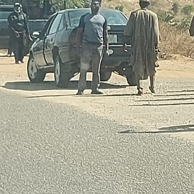 This image, captured in Barakin Kuru III, Nigeria, depicts an outdoor scene on an unpaved or gravel road during daylight. Three male individuals and two vehicles are present. In the mid-ground, a dark-colored sedan is stationary; its front driver's side door appears partially open. To the left of the sedan, a male individual stands, wearing dark clothing and a light-colored head covering. A second male individual stands in the central foreground, positioned slightly to the right of the sedan's rear. He wears a light blue short-sleeved shirt and dark trousers, facing towards the viewer, and holds a dark, possibly reddish, object under his left arm. A third male individual is observed from behind, walking towards the right side of the frame. He wears a long, light-colored traditional garment. Partially visible behind the sedan is a second dark-colored vehicle, consistent with an SUV or truck. The background consists of dry, sparse vegetation on a sloping terrain.