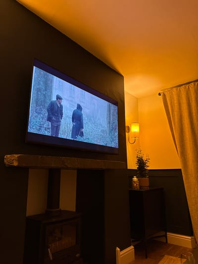 The image shows an interior view, focusing on a television mounted on a dark wall. The screen displays a scene with two men in dark, period clothing walking through a wintry landscape, with a building visible in the background. Above the television is a small, cylindrical lamp emitting a warm glow. A tan-colored curtain is visible at the top of the frame. To the right of the television, there's a dark shelving unit with decorative items, including greenery. A fireplace insert is present along the bottom. The setting appears to be indoors, and the overall lighting is subdued, contributing to a cozy atmosphere. While the location of the depicted scene in the city of Windermere, United Kingdom is not stated, it is likely that the room is decorated in a style that reflects the region's atmosphere and culture.