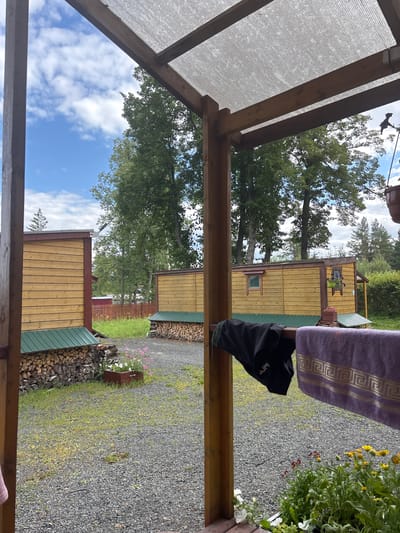 The image depicts an outdoor scene on a cloudy day, viewed from under a wooden and plastic-covered shelter. In the foreground, a wooden railing supports a black garment and a purple towel with a gold geometric pattern. Beyond the shelter, two yellow wooden structures, possibly cabins or small houses, are visible. The structures have green metal roofs. Piles of firewood are neatly stacked against the foundations of these buildings. A gravel driveway or yard surrounds the structures, with patches of green grass and some flowering plants. Tall green trees form a backdrop against the sky, which is partially filled with white clouds. The scene appears to be in a rural or semi-rural setting, consistent with the location in Shuiskaya Chupa, Russia.