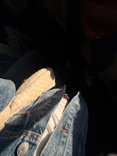 A cream-colored woven handbag rests against a denim jacket and jeans, with a glimpse of a white shirt underneath. A button is visible on the jacket, along with brown stitching. The scene is split by contrasting light and deep shadows. In the obscured areas are vague suggestions of a brown bag, a patterned item, and shoes.