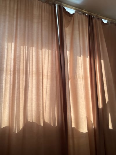 The image shows a close-up of sheer, beige curtains hanging indoors. Sunlight streams through the fabric, creating vertical stripes of light and shadow. The bottom edge of the curtains forms an uneven, scalloped silhouette against the illuminated background. The scene appears to be in an apartment in Novogorelovo, Russia, possibly a bedroom or living room, given the presence of decorative molding at the top of the window frame. There are no people or specific objects visible beyond the curtains and the implied window.