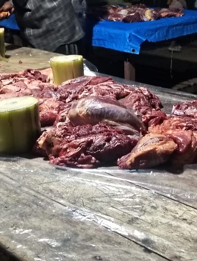 This media file depicts a scene at a market in Cottrueng, Indonesia, likely a butcher's stall. The primary subjects are large cuts of raw, red meat, piled on a wooden table covered with a plastic sheet. Two cylindrical, pale green objects, possibly stalks of banana tree, are placed near the meat. In the background, a person wearing a checkered shirt is partially visible, with their hands appearing to be engaged with the meat or preparing the stall. A blue tarp is spread over a surface in the background, suggesting a covered market area or vendor's setup. The lighting suggests it could be daytime or a well-lit indoor space. The overall impression is one of a busy, active marketplace where raw meat is being sold. There is no visible text in the image.