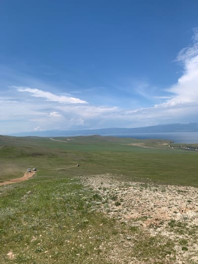 A wide-angle, eye-level shot captures a vast, rolling green landscape under a bright blue sky with scattered white clouds. The foreground is characterized by sparse, dry grass and rocky terrain. In the middle ground, a dirt road winds through lush green hills. Several white vans are visible on the road, with two clustered together on a bend and one further away. In the background, a wide body of water, likely Lake Baikal, stretches to the horizon, bordered by a distant, mountainous shoreline. A small settlement with some yellow structures is visible near the water's edge. The overall scene depicts a remote, natural landscape in Malyi Khuzhir, Russia.