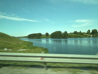 A landscape photograph captures a serene view of a body of water, likely a river or lake, under a clear, pale blue sky with scattered wispy clouds. The water's surface is calm, reflecting the muted colors of the sky and the surrounding greenery. On the left side of the frame, a grassy hill slopes down towards the water, dotted with patches of low-lying vegetation and some scattered stones. The right bank of the water is lined with a dense fringe of trees, their foliage a deep green. Further in the distance, more trees and a few small buildings can be seen on the horizon. In the foreground, a gray metal barrier, possibly a guardrail, stretches horizontally across the lower portion of the image, obscuring the very bottom of the scene and suggesting the photograph was taken from a moving vehicle. The overall impression is one of natural beauty and tranquility in a rural setting.