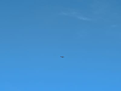 An airplane is visible in the sky above Peetri alevik, Estonia. The sky is clear and blue, with a few wisps of clouds. The airplane is small in the frame, appearing as a dark silhouette against the bright sky. It is flying horizontally, with its wings extended. The image appears to be taken from the ground, looking up.