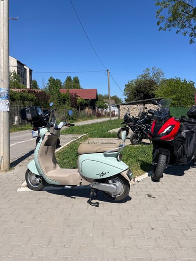 The image depicts a street scene in Petrovaradin, Serbia, featuring several parked motorcycles and scooters. In the foreground, there is a light-blue and beige electric scooter parked on a paved sidewalk. Behind it are two larger motorcycles, one with a red front fairing and black luggage. The street runs along the side of the sidewalk. Behind the scooters and motorcycles is a mix of residential buildings and greenery, with houses visible among the trees. A utility pole is present near the foreground. The sky is clear and blue, suggesting a sunny day. The license plate is visible on one of the scooters, indicating local registration.
