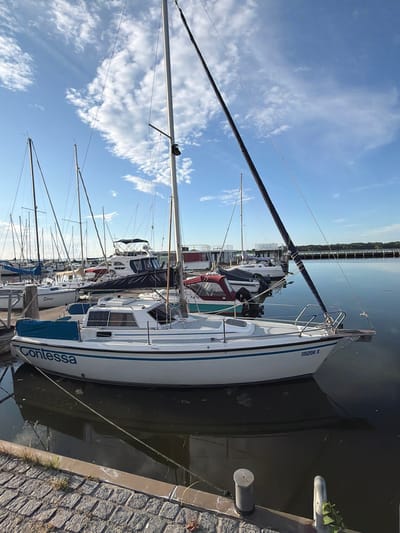 The image shows a marina in Ribnitz-Damgarten, Germany. The focus is on a white sailboat named "Contessa" docked at the marina. The boat has a blue stripe along its hull and a registration number "115209 S". Other sailboats and a larger motorboat are visible in the background, as well as a pier and buildings on the shore. The sky is blue with white clouds, and the water is calm, reflecting the boats and the sky. The foreground shows a cobblestone walkway and mooring bollards. There are no people visible in the image.