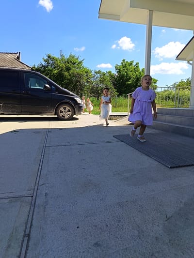 In Egorovca, Moldova, on a sunny day, a black van is parked on a concrete driveway. Several young girls in dresses are running towards a building with a covered porch and stairs. One girl in a purple checkered dress is in the foreground, looking to her left with her mouth open. Another girl in a white dress is behind her, further to the left. In the background, more children are visible, along with trees and a cloudy blue sky. The scene appears to be a residential area.