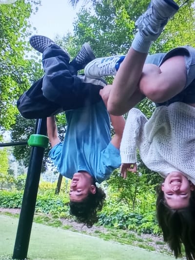 Two young individuals are depicted hanging upside down at an outdoor recreational area in what is identified as Kasagaran, Uzbekistan. The scene is captured from a low angle, looking upwards towards the subjects and a bright, overcast sky.
The individual on the left, a male in his late teens or early twenties, has dark, curly hair hanging downwards. He is wearing a light blue t-shirt, dark pants, and black and white checkerboard-patterned athletic shoes. His knees are hooked over a horizontal bar, which is supported by a black vertical pole with a green plastic grip near its top. His facial expression is neutral to slightly smiling.
The individual on the right, a female in her late teens or early twenties, has dark, straight hair also hanging downwards. She is wearing a light-colored, possibly off-white, knitted long-sleeved sweater, light-colored shorts, white socks, and white athletic shoes with blue accents. She is also inverted, positioned closely to the male subject, with her legs appearing to be intertwined or supported by his stance on the bar. Her face is visible upside down, showing a broad smile.
The environment includes lush green foliage, likely trees and bushes, in the background, suggesting a park or similar green space. The ground beneath the subjects features green artificial turf. The overall lighting suggests natural daylight.