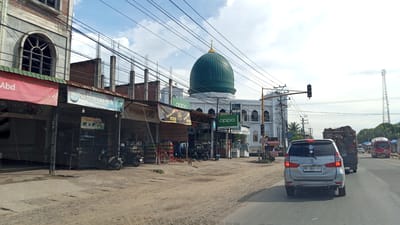 The image depicts a street scene in Cotpuuk, Indonesia. A silver minivan with license plate BK 1843 UT is in the foreground, driving on a paved road. Behind it, a truck loaded with what appears to be lumber or cardboard is also moving forward. To the left of the road, a row of shops lines the street, displaying various signs, including "SEJAHTRA" in red and white, and a store with "IKAN BAKAR" (grilled fish) advertised. A mosque with a prominent green dome and a crescent moon is visible in the background, situated behind some of the shops. The sky is partly cloudy. A traffic light is visible on the right side of the road. Utility poles and wires are strung across the scene. A cell tower is in the distance to the right.