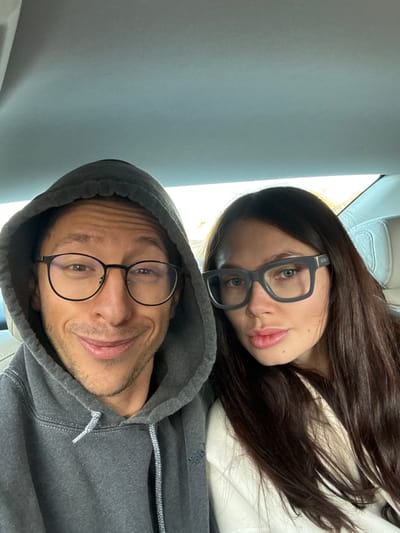 A medium close-up photograph displays two individuals, one male and one female, seated within a vehicle. The male, positioned on the left, wears a grey hooded sweatshirt with the hood up, revealing an 'adidas' logo on the chest, and dark-framed round eyeglasses. He looks directly at the camera with a slight smirk. The female, to his right, has long dark hair and wears large rectangular grey eyeglasses with 'GUCCI' visible on the right temple. She presents a neutral expression with slightly pursed lips, wearing a light-colored top. The vehicle's interior shows a light-colored headliner and beige seat upholstery behind the female. Bright daylight is visible through a rear window, creating a backlit effect. The scene is located in the city of Ramat Negev Regional Council, Israel.