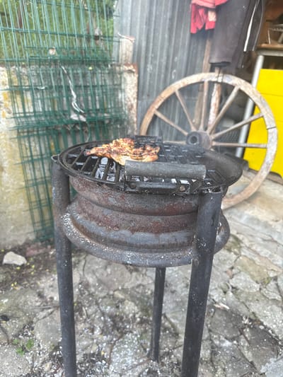 The image depicts a piece of grilled meat cooking on a rustic, black metal grill. The grill is positioned outdoors on a concrete or stone surface, next to a green wire fence and a wooden wagon wheel. The surrounding environment shows signs of wear and age, suggesting a backyard or outdoor cooking area in the countryside. The location in the city of Boltata, Bulgaria is noted, but no elements in the image directly confirm this. There are no people visible in the frame.
