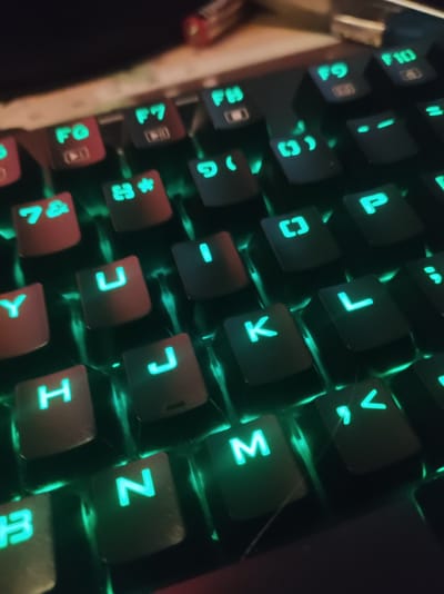A close-up, high-angle shot shows a backlit keyboard with green illumination. Keycaps display a variety of characters, including letters, numbers, and function keys. Some keycaps are red, while others are black. The keyboard appears to be a standard QWERTY layout, with some keys featuring additional symbols or icons, like the F6 and F7 keys, which display play and record icons. The lighting creates a glow around each illuminated character. The background is dark and out of focus, with some hints of metallic objects. No people or city elements are visible in the image.