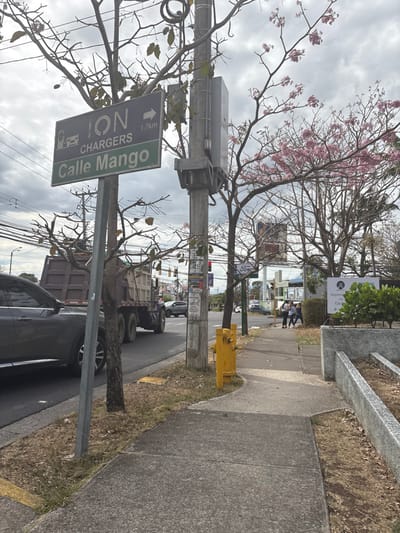 The image shows a street corner in San Rafael, Costa Rica, located at latitude 9.937, longitude -84.152. A street sign reads "ION CHARGERS Calle Mango 1.7km" and points to the right.  A grey car is parked on the left side of the street, a large truck is stopped on the right side of the street, and there is a yellow fire hydrant on the sidewalk to the right of the street sign. A group of three people walk on the sidewalk in the distance, heading toward the right.  The street is lined with trees with pink blossoms, and there are several other street signs and electrical equipment on a utility pole. There is a small, leafy bush in the foreground of the image, next to the street sign. The sidewalk is cracked and worn.