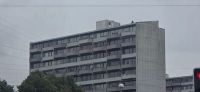 This is a media file depicting an overcast day in Aarhus, Denmark. The primary subject is a large, modern apartment building with a grey facade and numerous balconies. The building is several stories high, with a repeating pattern of windows and cantilevered balconies. Some balconies have railings, while others appear to be open. A portion of another building, also with a similar architectural style, is visible to the right.
Several power lines are strung across the sky in the foreground, creating a layered effect against the building and the sky. The sky is uniformly grey, indicating cloudy or overcast weather, and there is no direct sunlight. To the left of the main building, the tops of green trees are visible, suggesting a leafy or park-like area nearby.
On the far right, a traffic light is partially visible, showing a red signal. This implies the presence of a road or intersection. The overall scene is urban and functional, typical of a residential or mixed-use area in a European city. There are no people or specific activities depicted, focusing instead on the architecture and the urban environment under a muted sky. The concrete facade of the main building on the right side has vertical lines, adding a textural element to its surface. The time of day is indeterminate due to the overcast conditions, but it appears to be daytime.
