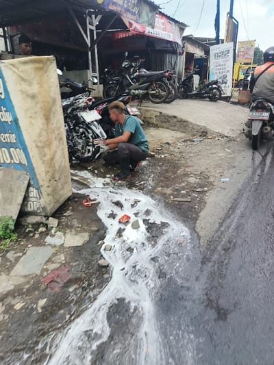 A man squats washing a motorcycle with soapy water, which is running onto the street. Additional motorcycles are parked nearby under a shaded awning of a business. A sign partially obscures the man, advertising "alju emir 10.000." Another sign in the background is for "BAN MOTOR SERVIS GRATIS." A person on a motorcycle is on the street, which appears damp.