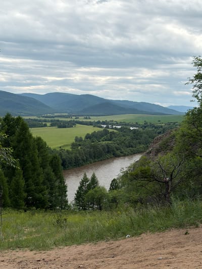 The image depicts a scenic natural landscape with a wide river flowing through a valley. The river, with its muddy brown water, curves through a lush green expanse dotted with trees. The valley is framed by rolling hills and distant mountains, their slopes covered in a mix of forests and fields. The sky above is overcast with a layer of grey clouds, suggesting a diffused light. In the foreground, a sandy embankment slopes upwards, covered with dry grass and scattered small stones. To the left, a dense cluster of tall evergreen trees dominates the view, while to the right, a rocky outcrop is partially covered with vegetation and a few bare branches. The scene is one of a rural, undeveloped environment, likely in a mountainous region. The location is identified as Ulekchin, Russia. There are no people or specific actions depicted in the image.