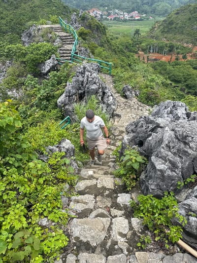 A man walks down a stone staircase on a rocky hillside. He is wearing a light-colored t-shirt, khaki shorts, a dark baseball cap, and gray sneakers. He is holding a dark object in his right hand. The staircase is bordered by lush green foliage and large gray rocks. In the background, a valley opens up with a village and green fields. The scene is set in the city of Tam Thanh Ward, Vietnam, suggesting a natural landscape with signs of human presence and development.
