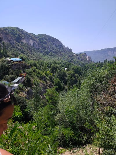 The image captures a panoramic view of a mountainous landscape in Arstanbap, Kyrgyzstan, dominated by lush green trees covering the slopes. In the mid-ground, on a rocky outcrop, several structures with blue and green roofs are visible, suggesting a recreational area or small buildings. A prominent zipline stretches across the frame, with one gondola suspended mid-air. The foreground is partially occupied by the shoulder and back of a person wearing sunglasses, looking down towards the scenic vista. The sky is clear and blue, indicating a sunny day. The overall scene depicts a natural environment with tourist amenities, possibly an adventure park or a viewpoint.