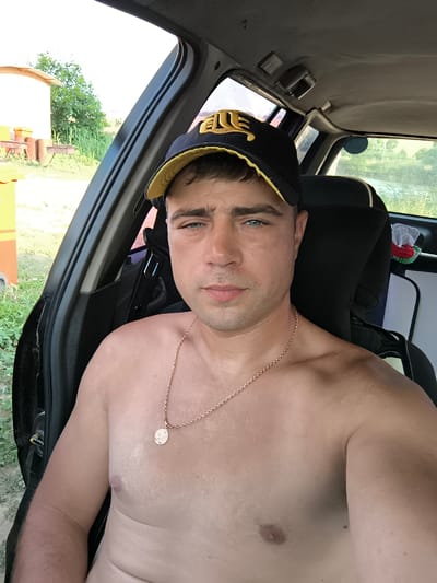 A shirtless man wearing a black and yellow baseball cap sits in the driver's seat of a car. He has a gold chain with a round pendant around his neck. The car appears to be parked outdoors, with greenery and a small building visible in the background. The lighting suggests it is daytime.